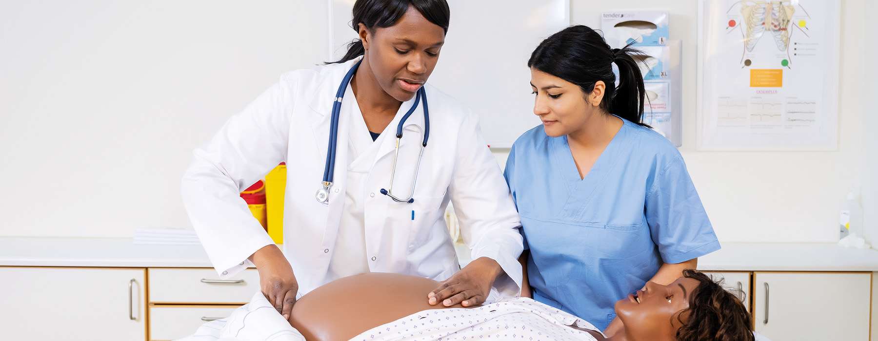 A healthcare professional in a white coat and a student in blue scrubs practice a physical abdominal examination on a pregnant patient simulator.