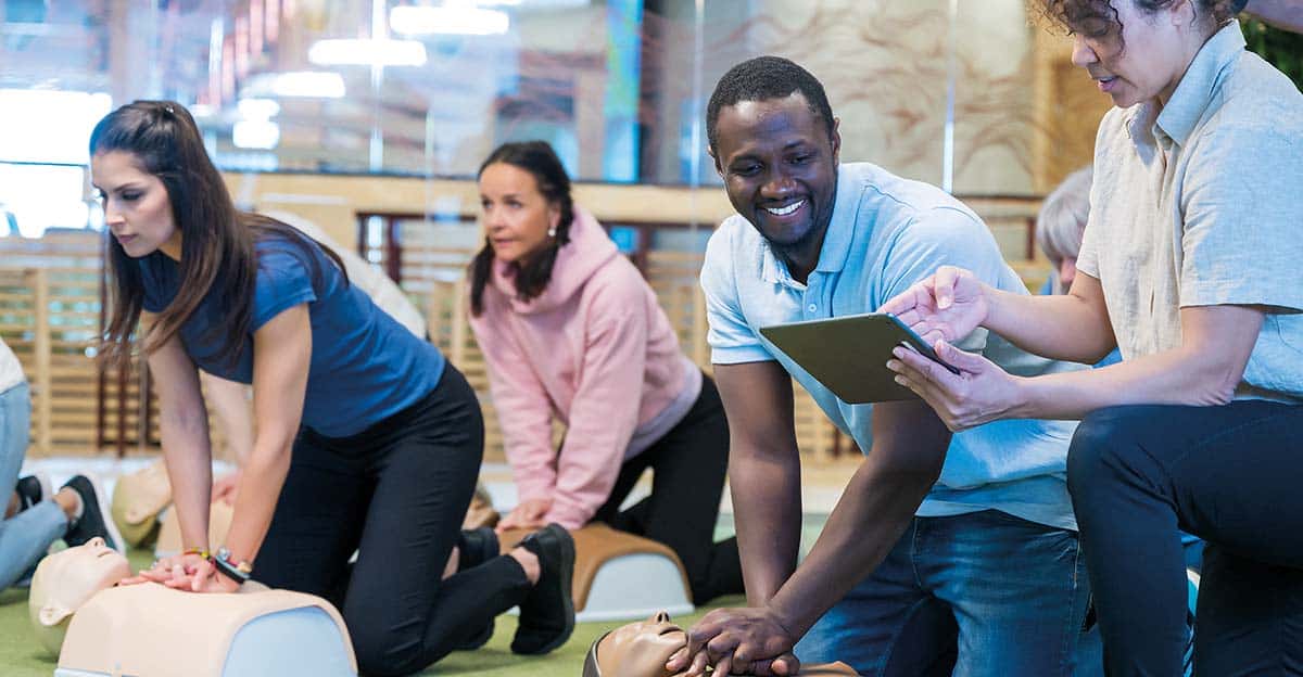 students practicing chest compressions on mannequins while an instructor provides guidance using a tablet in a classroom setting