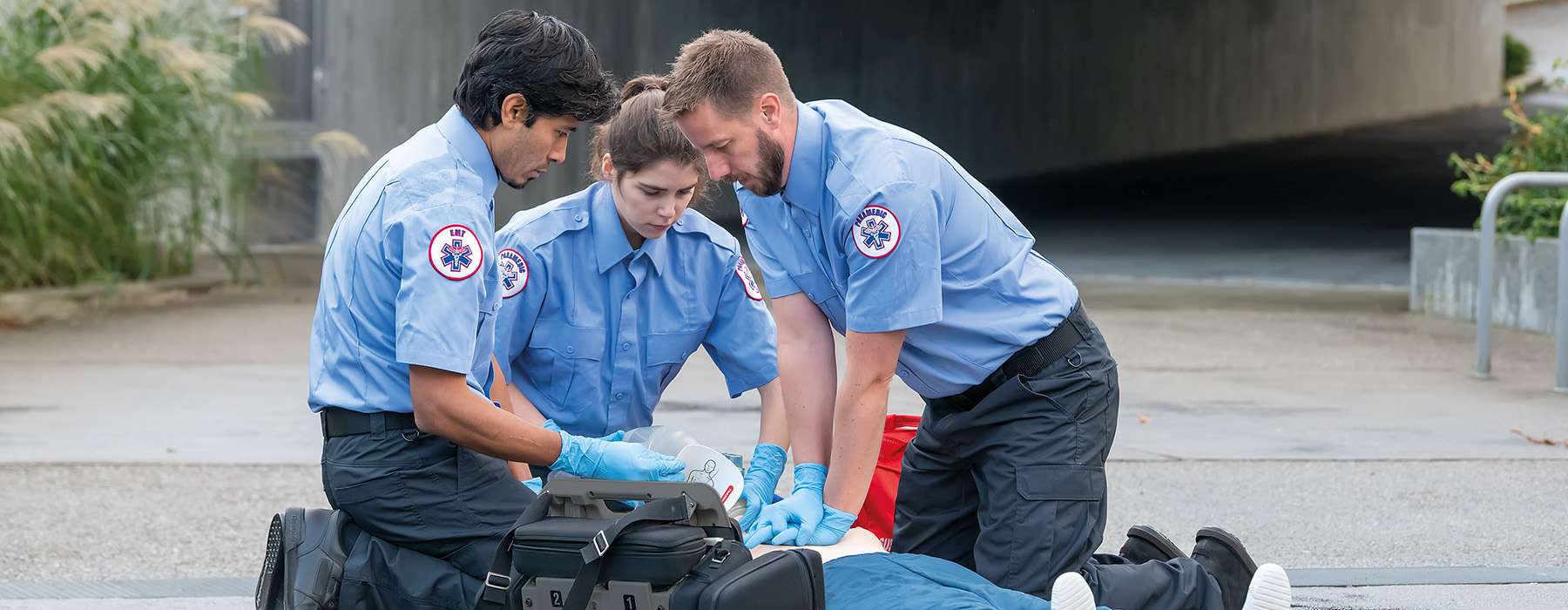 A team of three EMS professionals practicing life-saving resuscitation and airway management on a simulator during an outdoor training session.