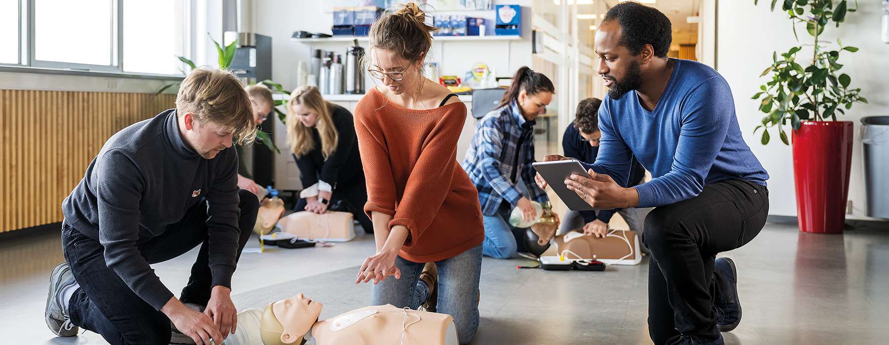 Several people in a bright, modern office environment practicing emergency medical procedures on various simulators under supervision.