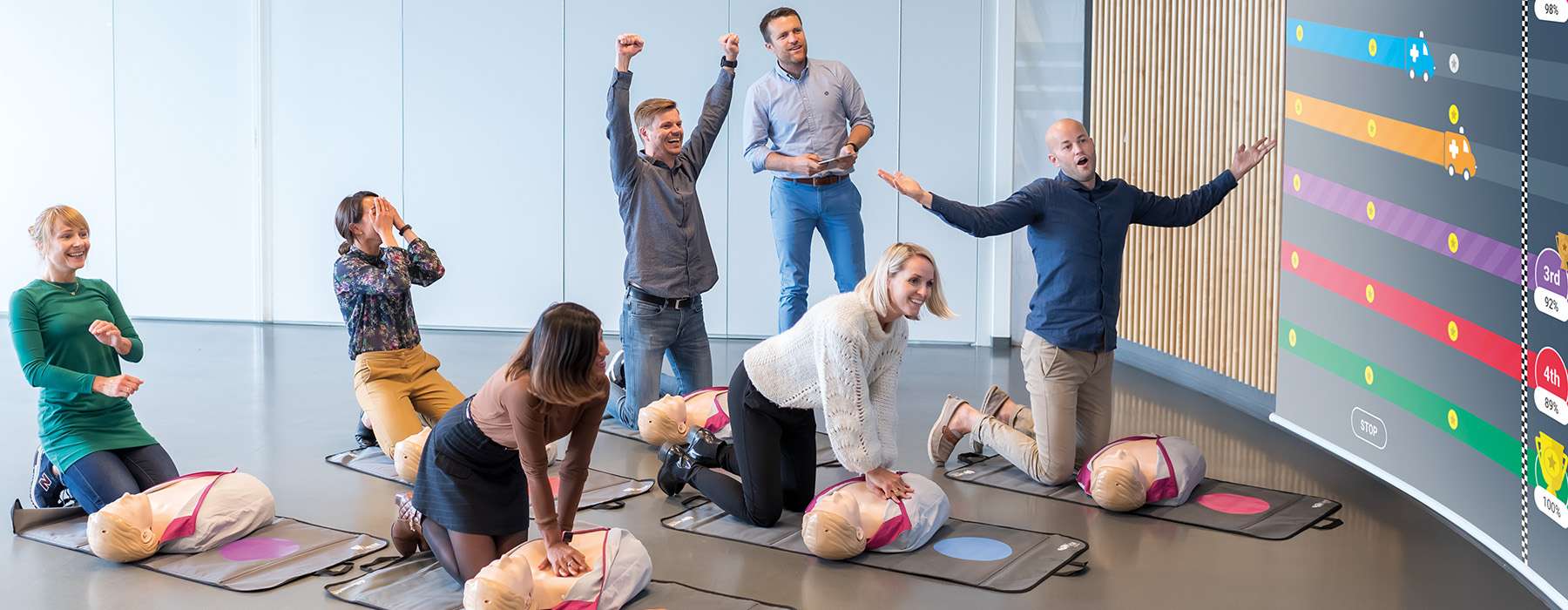 A group of people cheering while practicing CPR on simulators, with a leaderboard displayed on a large screen in the background.