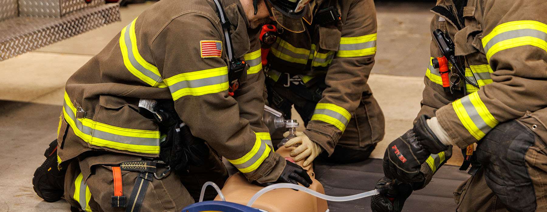 Three firefighters in full protective gear practicing life-saving resuscitation and airway skills on a manikin.