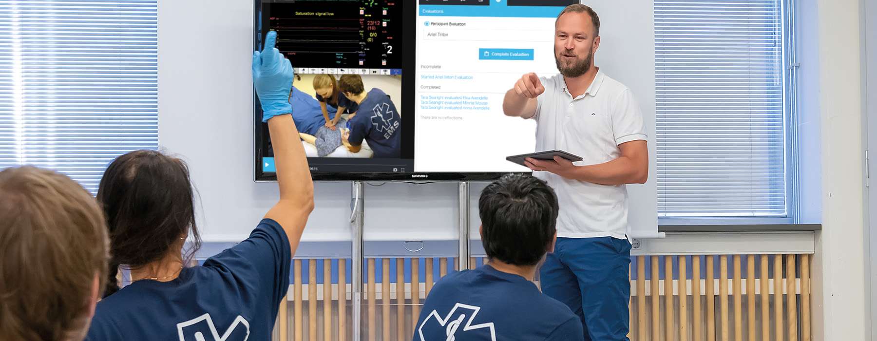 An instructor standing in front of a large screen, using a tablet to evaluate a team's performance during a recorded simulation session.