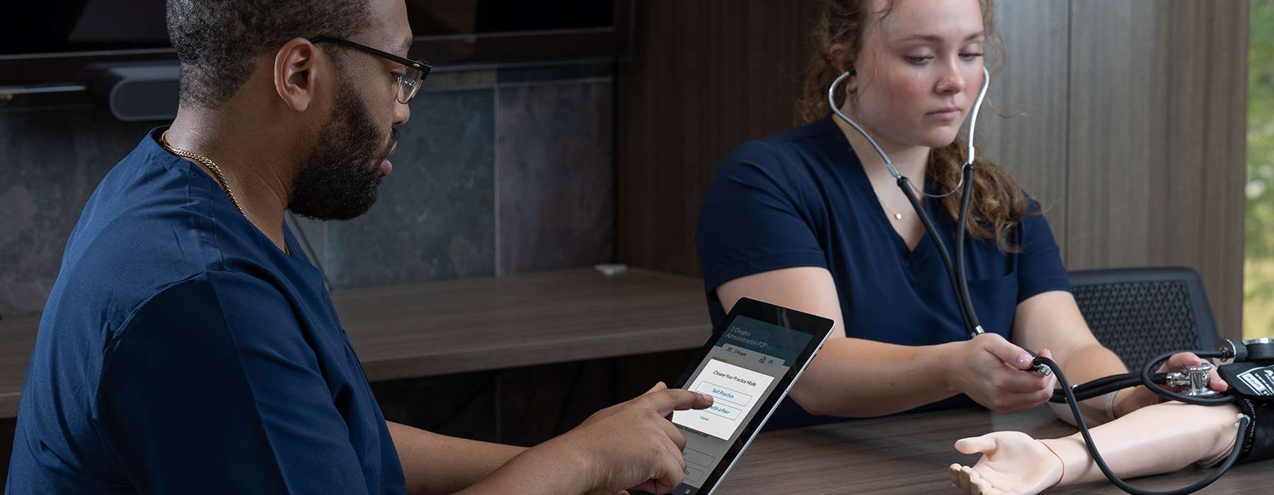 A student uses a tablet-based learning application to guide a peer through a physical assessment and stethoscope skills drill.