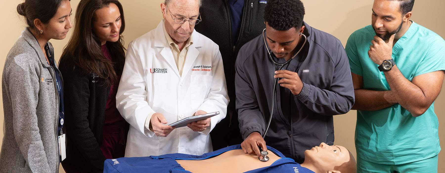 A medical team, including a senior doctor with a tablet, observes a student practicing heart sound auscultation on a cardiology simulator.
