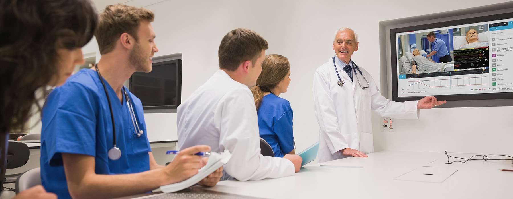A clinical instructor in a white coat uses a large screen to lead a simulation debriefing session, reviewing recorded footage and event logs for medical students.