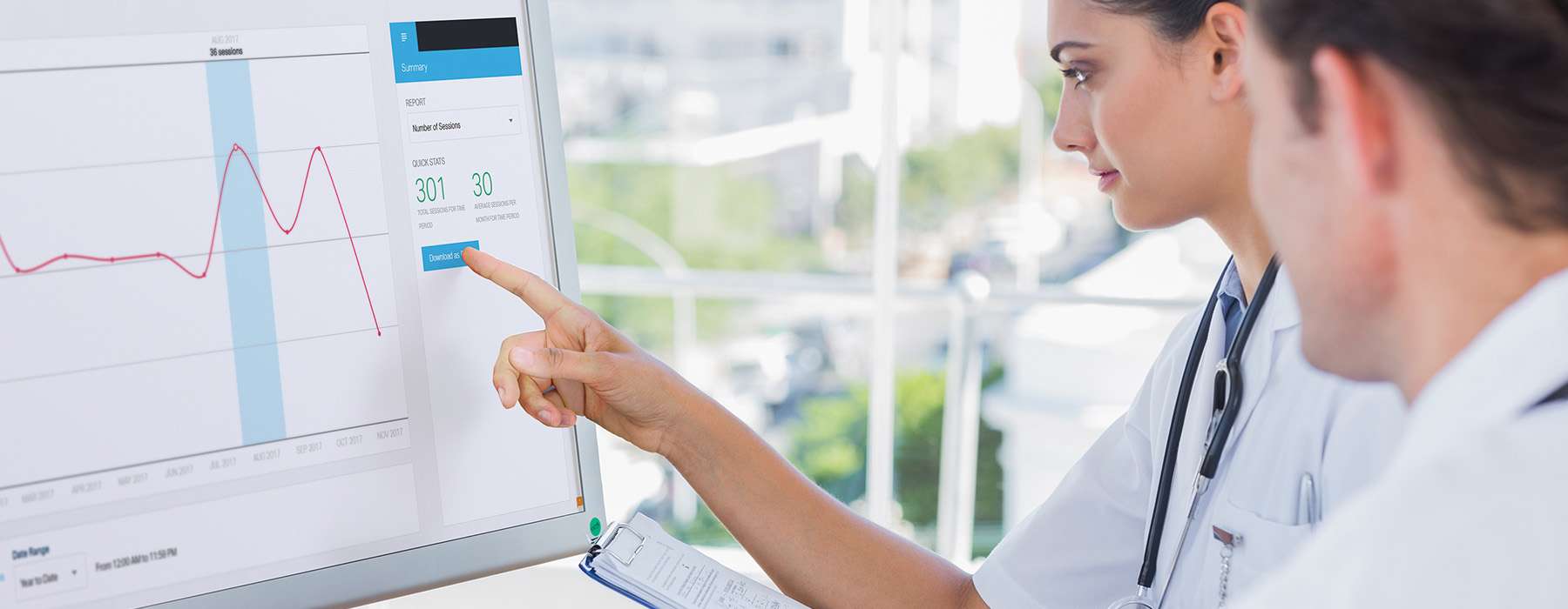 Two healthcare professionals in lab coats review digital performance metrics, session statistics, and trend charts on a large monitor to evaluate training results.