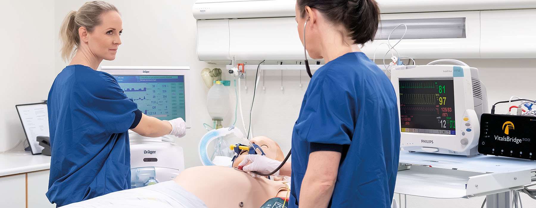 Two healthcare professionals monitoring vitals and using a stethoscope on a patient simulator equipped with advanced medical monitors and a VitalsBridge unit.