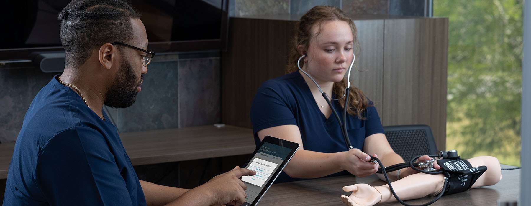 A healthcare student uses a tablet-based learning application to guide a peer through a blood pressure assessment on a simulator arm.