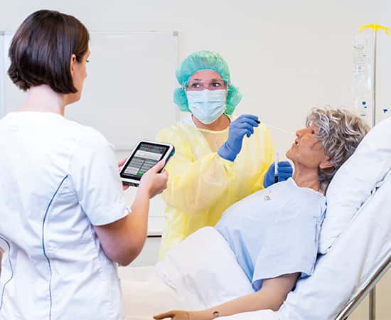 Two nurses, one wearing a mask and other protective gear, training using the Nursing Anne Simulator