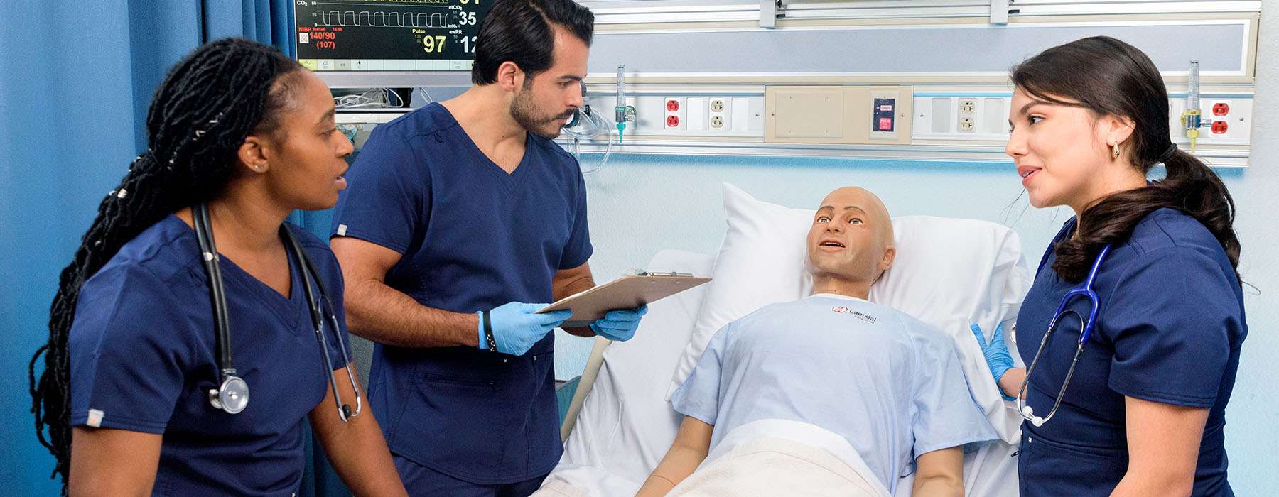 Medical team standing around a hospital bed occupied by a male Nursing Anne simulator during training.