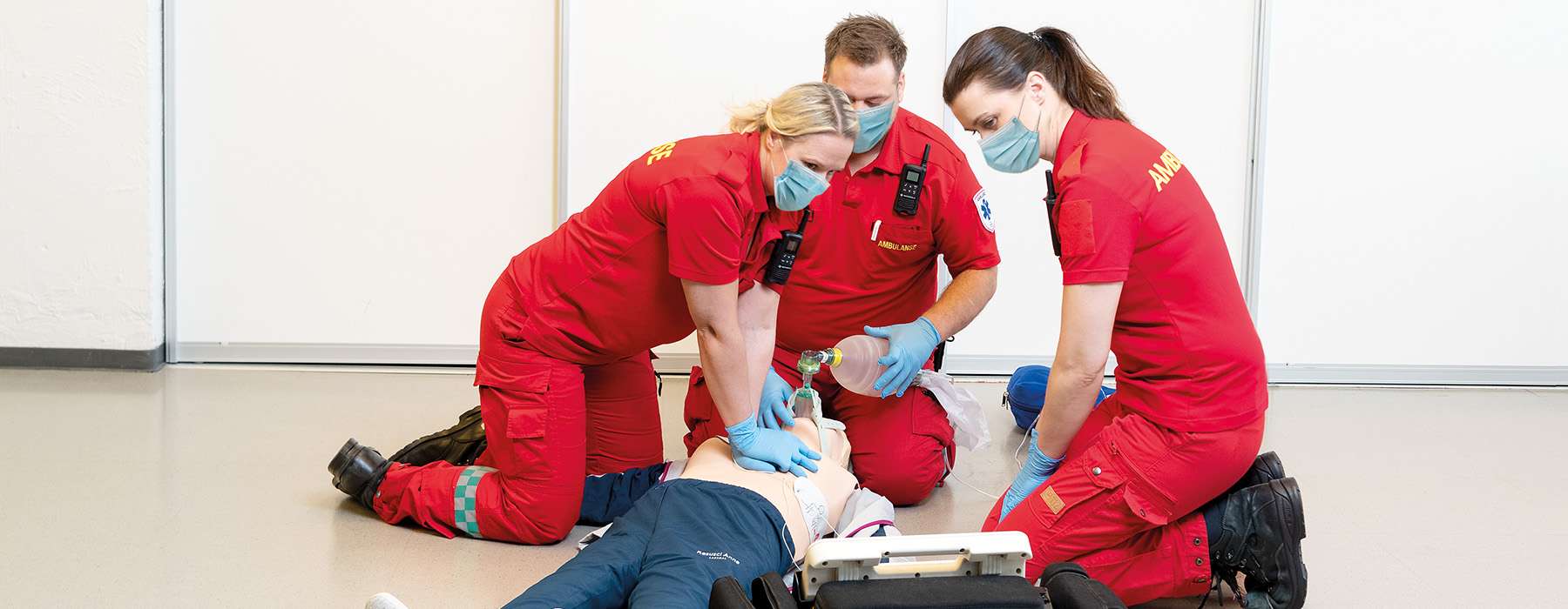 3 EMS workers in red clothing doing CPR training on a manikin.