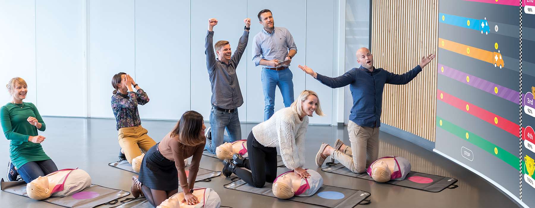 Group of people doing CPR training on manikins as a competition, while their scores show as a score on a screen.