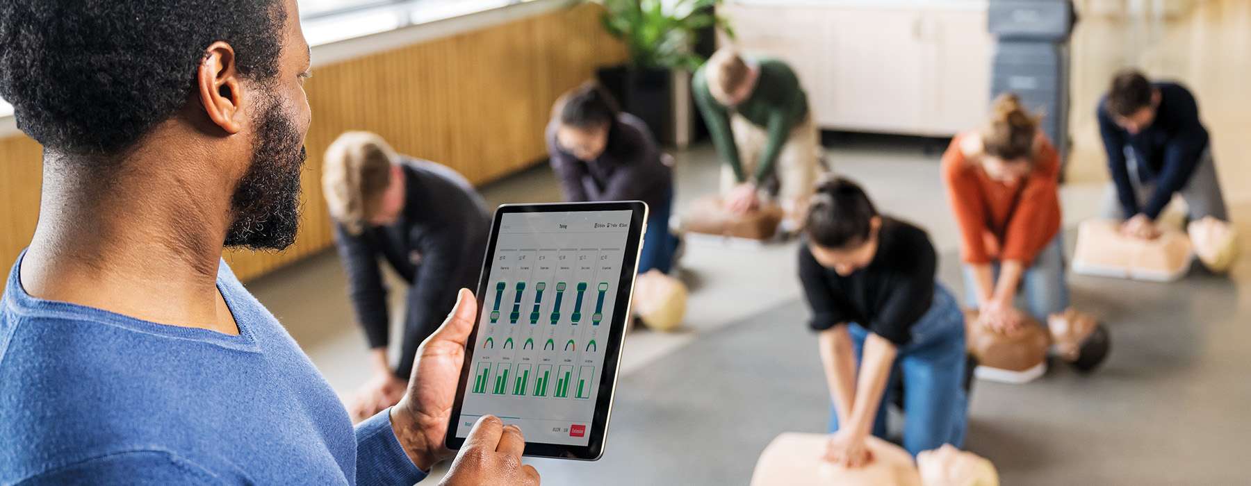 Man in foreground watching a group of people doing CPR training on manikins on the floor. The man is holding a tablet and following stats.