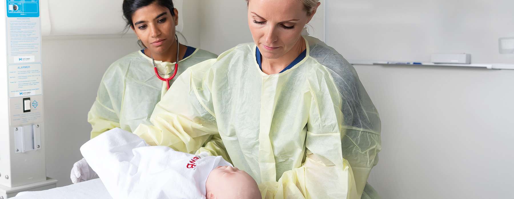 Two nurses wearing yellow operating clothes. One of them is holding a newborn manikin.