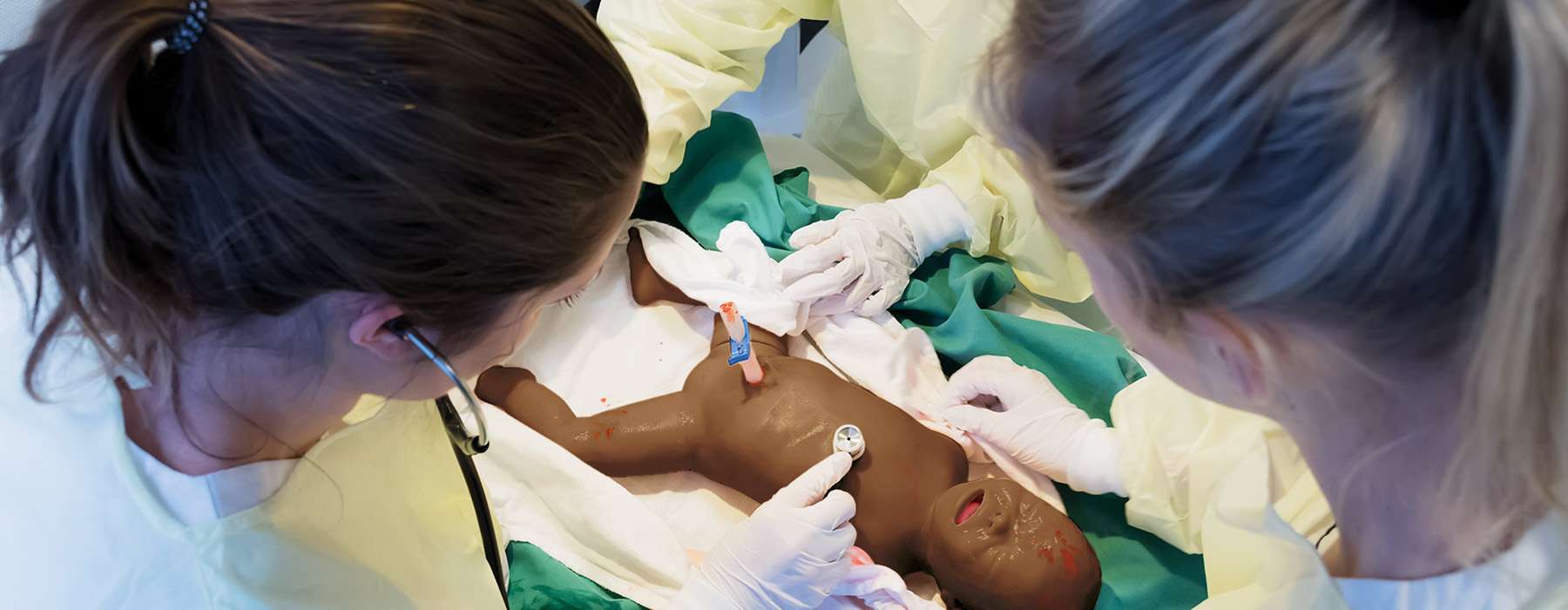 A top-down view of two healthcare providers in protective gear using stethoscopes to assess an infant patient simulator during high-fidelity training.