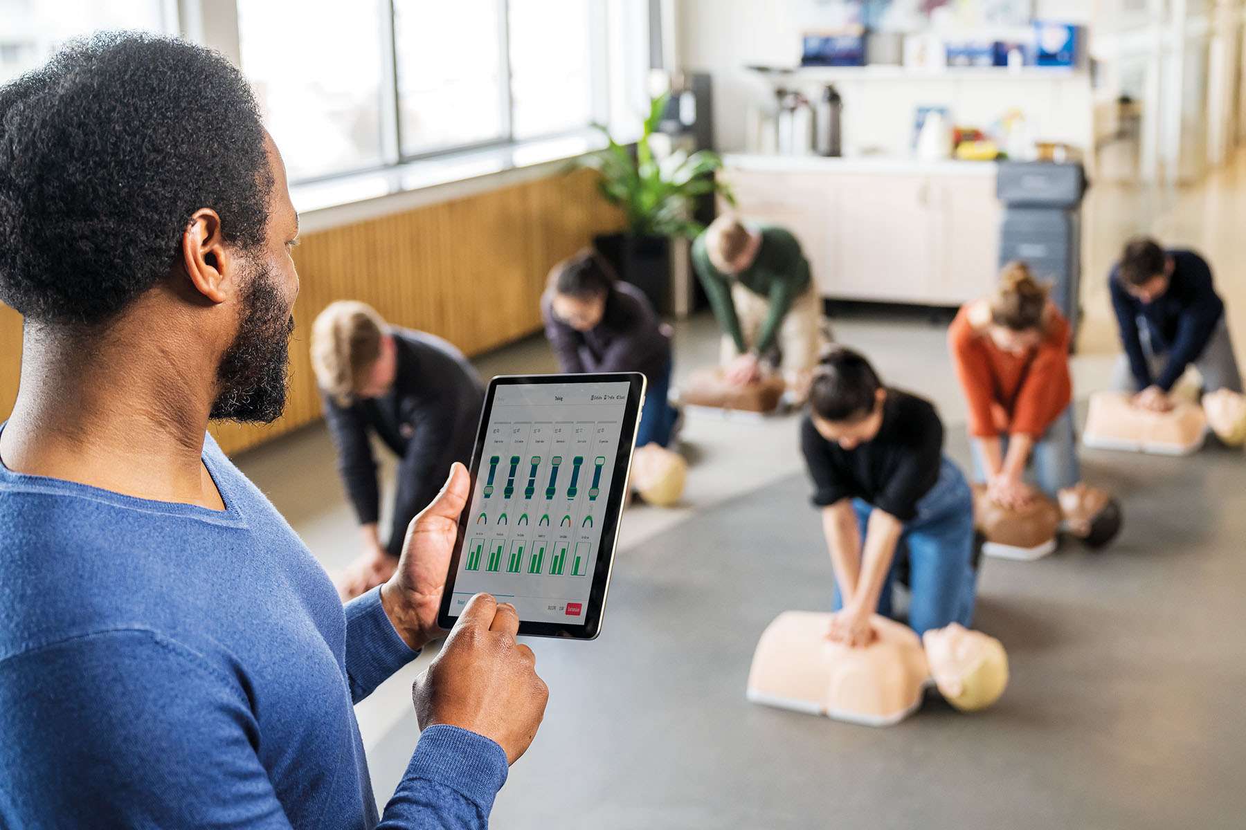 man reviewing stats on a tablet while watching people do CPR training in front of him.