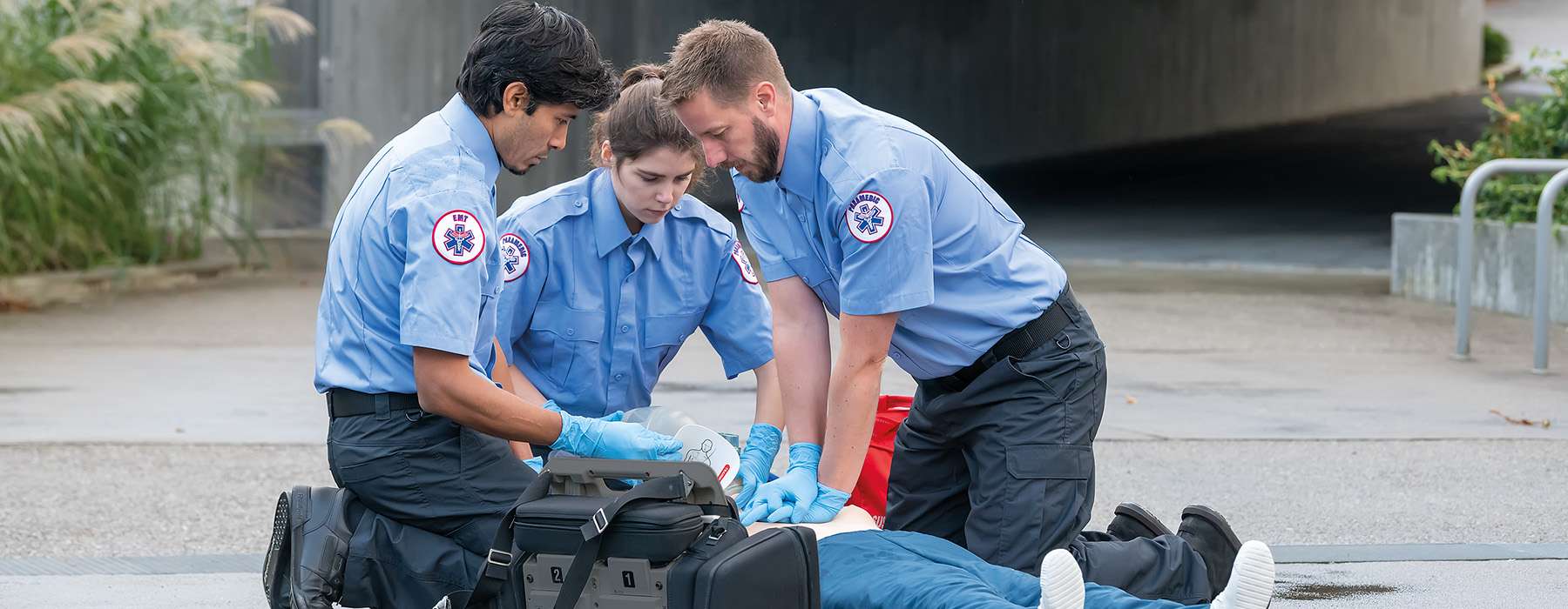 Three Emergency Medical Services (EMS) trainees or workers practicing high-quality chest compressions and ventilation on a manikin during an outdoor training session.