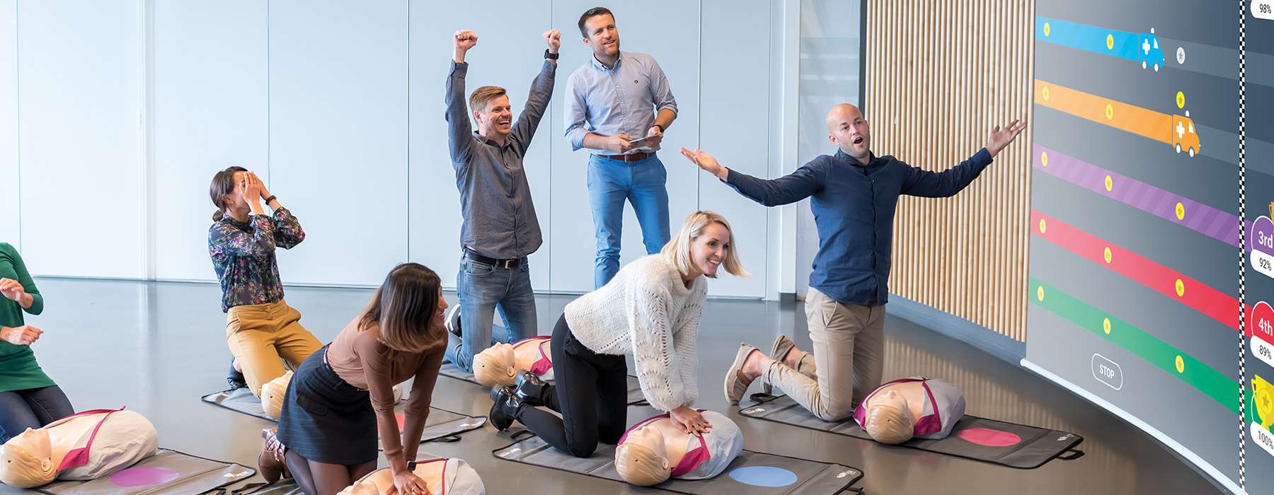 A group of people performing CPR on manikins in a classroom setting, with visual feedback from a large digital screen showcasing gamified real-time performance data for competitive training.