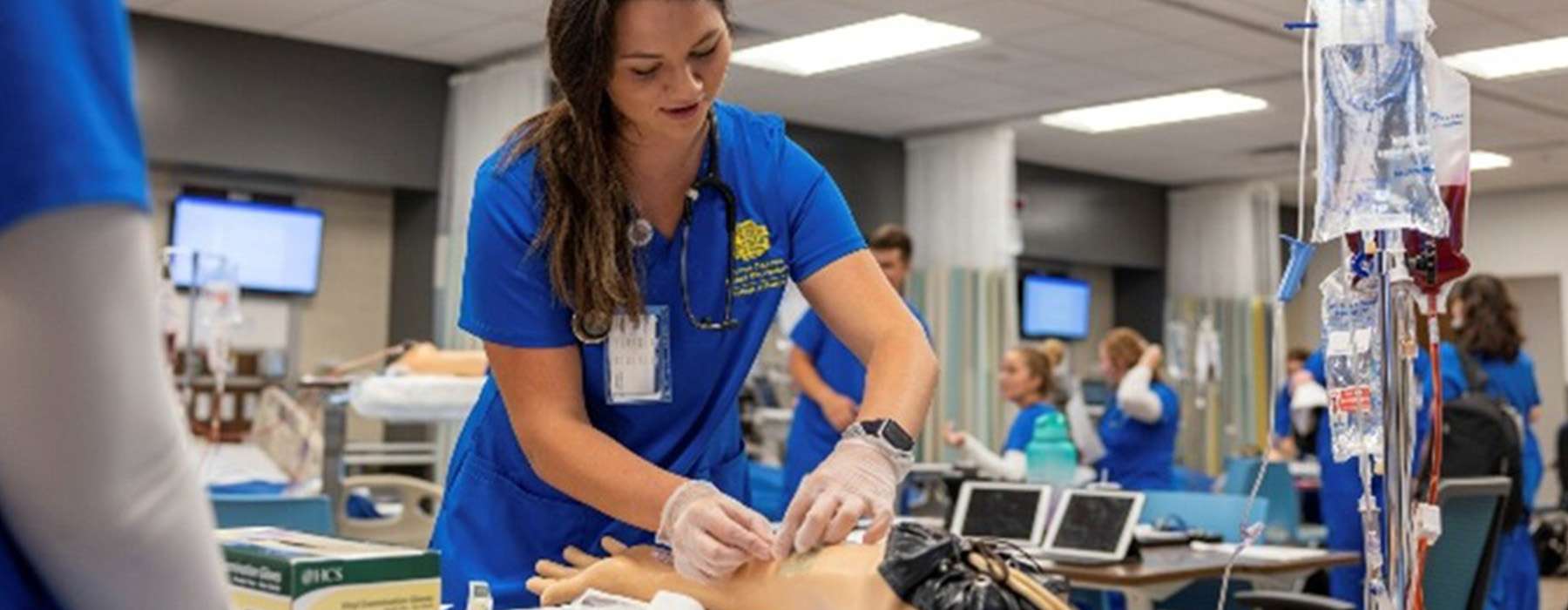 A nursing student practicing medical procedures on an arm simulator in a training environment.