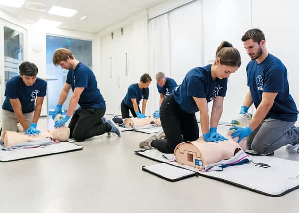 A group of six healthcare trainees or students practicing CPR skills on individual manikins in a bright classroom environment.
